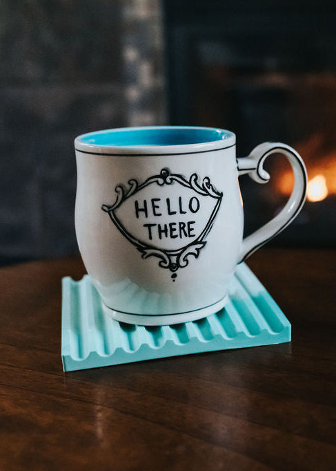 White mug with 'Hello There' text on a teal coaster, next to colorful coasters on a wooden surface with a fireplace in the background.